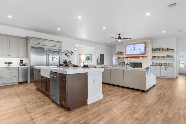 kitchen featuring backsplash, ceiling fan, a center island with sink, light hardwood / wood-style flooring, and a stone fireplace