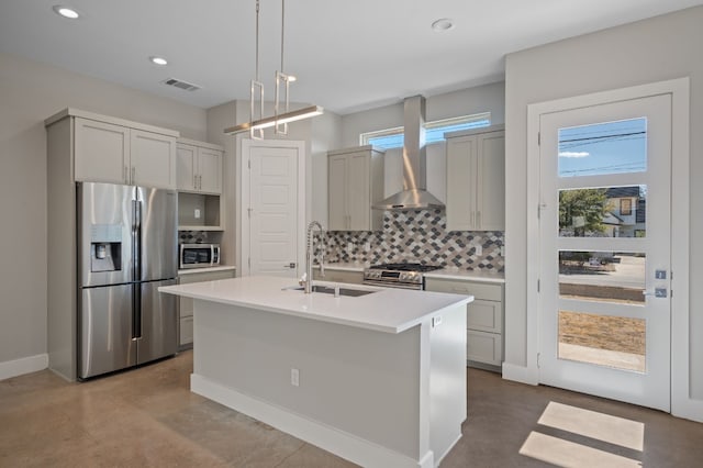 kitchen with sink, stainless steel appliances, wall chimney range hood, decorative light fixtures, and a kitchen island with sink