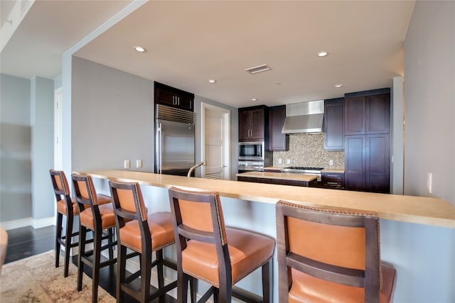 kitchen featuring a kitchen bar, built in appliances, tasteful backsplash, and wall chimney exhaust hood