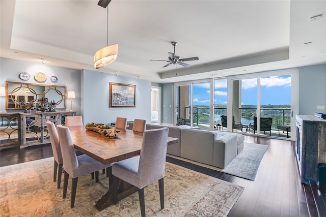 dining area with wood-type flooring, a water view, ceiling fan, and a tray ceiling