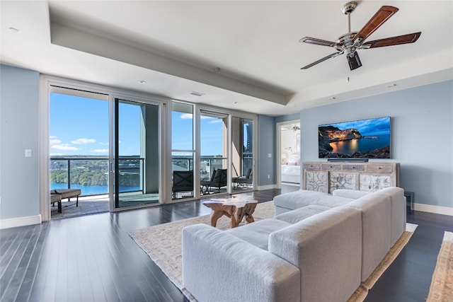 living room with a water view, a raised ceiling, dark wood-type flooring, ceiling fan, and expansive windows