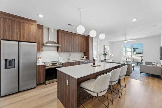 kitchen with stainless steel appliances, sink, wall chimney range hood, pendant lighting, and light hardwood / wood-style flooring