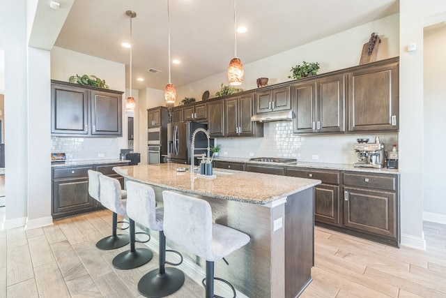 kitchen with tasteful backsplash, light stone counters, pendant lighting, a center island with sink, and appliances with stainless steel finishes