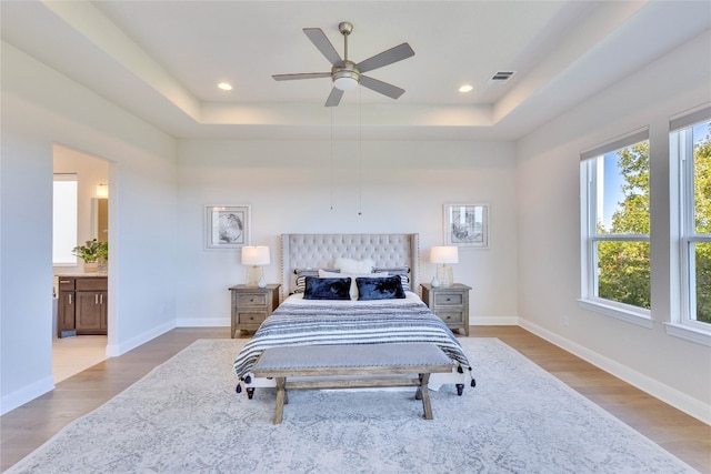 bedroom featuring a raised ceiling, ceiling fan, ensuite bathroom, and light wood-type flooring