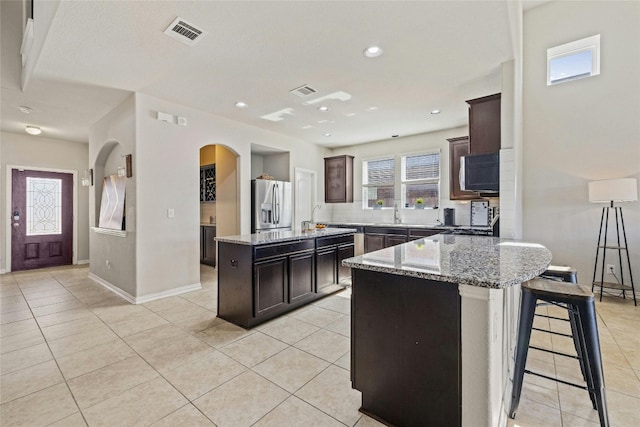 kitchen with light tile patterned flooring, stainless steel fridge, a kitchen island, light stone counters, and dark brown cabinetry