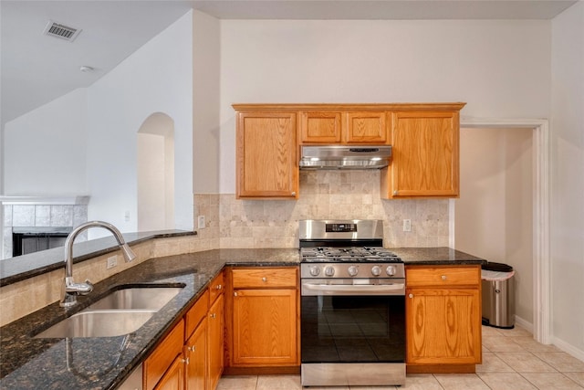 kitchen featuring a tiled fireplace, a sink, dark stone countertops, under cabinet range hood, and stainless steel gas range oven