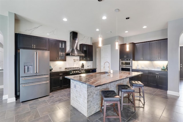 kitchen featuring sink, wall chimney exhaust hood, an island with sink, appliances with stainless steel finishes, and a breakfast bar area