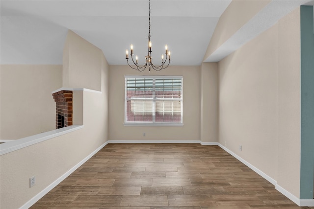 unfurnished dining area with a brick fireplace, an inviting chandelier, wood-type flooring, and vaulted ceiling