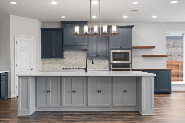 kitchen featuring a kitchen island with sink, hanging light fixtures, built in microwave, and stainless steel oven