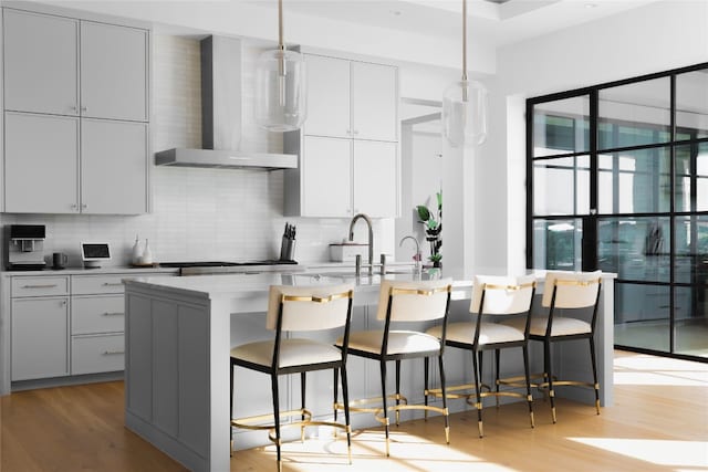 kitchen with a kitchen island with sink, wall chimney exhaust hood, hanging light fixtures, and light wood-type flooring