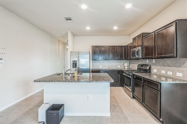 kitchen with appliances with stainless steel finishes, backsplash, dark brown cabinetry, an island with sink, and dark stone counters