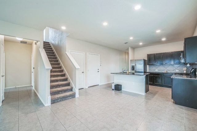 kitchen with decorative backsplash, dark stone counters, a kitchen island with sink, light tile patterned floors, and stainless steel refrigerator with ice dispenser