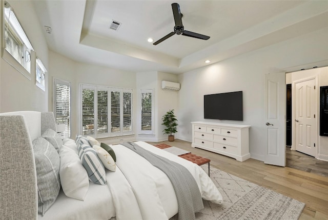 bedroom featuring a wall mounted air conditioner, light hardwood / wood-style flooring, a raised ceiling, and ceiling fan