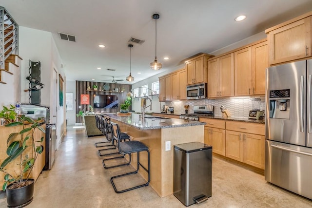 kitchen with ceiling fan, dark stone countertops, pendant lighting, a kitchen island with sink, and appliances with stainless steel finishes