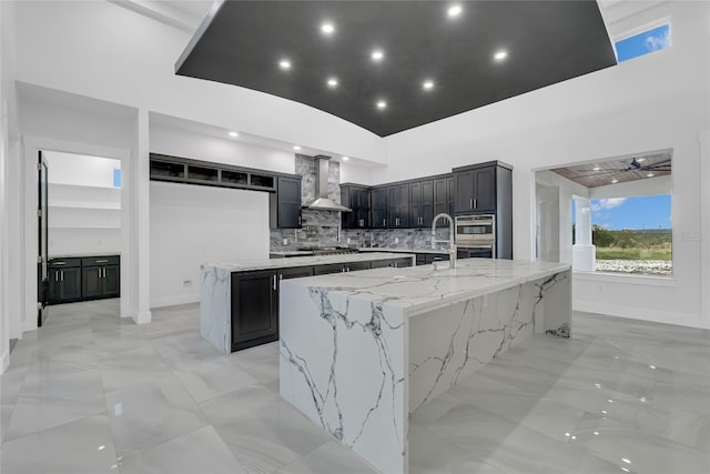 kitchen with decorative backsplash, appliances with stainless steel finishes, light stone counters, wall chimney range hood, and a large island