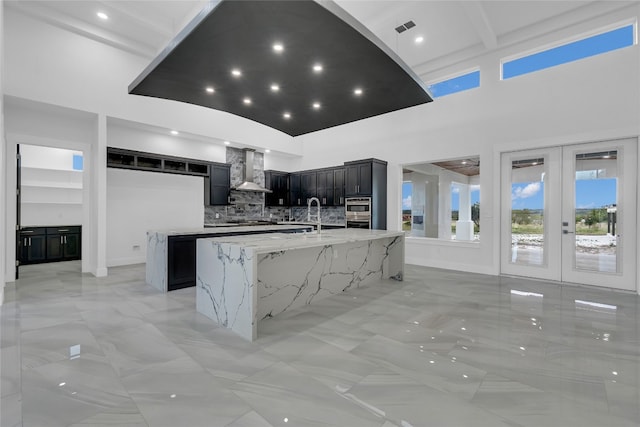 kitchen featuring french doors, a towering ceiling, tasteful backsplash, wall chimney exhaust hood, and a large island