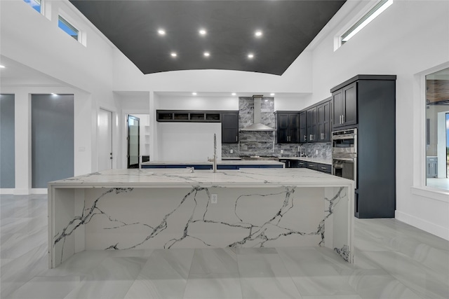 kitchen with light stone countertops, wall chimney range hood, backsplash, double oven, and a spacious island