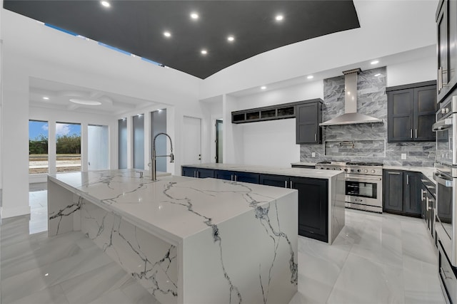 kitchen featuring light stone countertops, tasteful backsplash, stainless steel range, wall chimney range hood, and a large island with sink