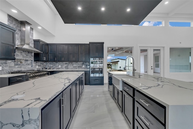 kitchen featuring wall chimney range hood, sink, decorative backsplash, light stone countertops, and a towering ceiling