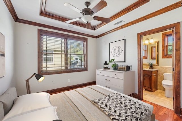 bedroom featuring light wood-type flooring, ensuite bath, ornamental molding, a tray ceiling, and ceiling fan