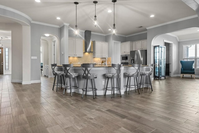 kitchen with stainless steel appliances, wall chimney range hood, crown molding, hardwood / wood-style floors, and white cabinets