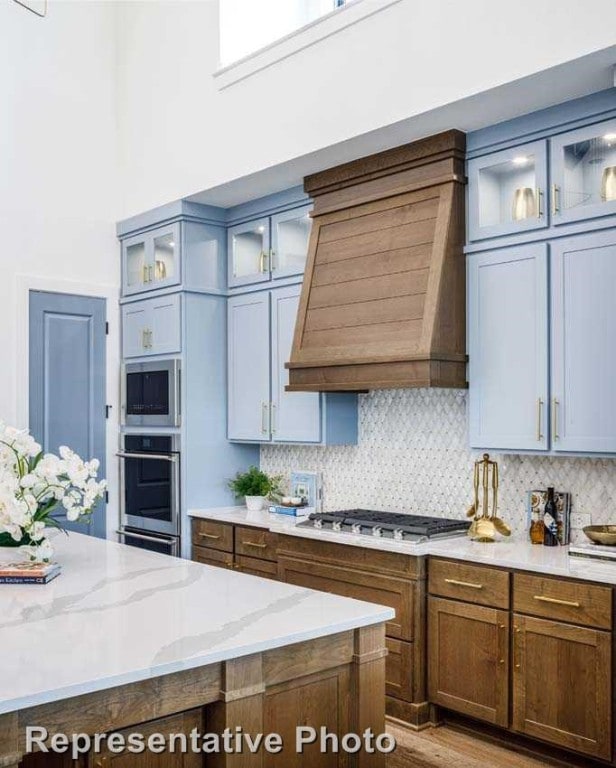 kitchen with light stone countertops, appliances with stainless steel finishes, tasteful backsplash, custom range hood, and light hardwood / wood-style floors