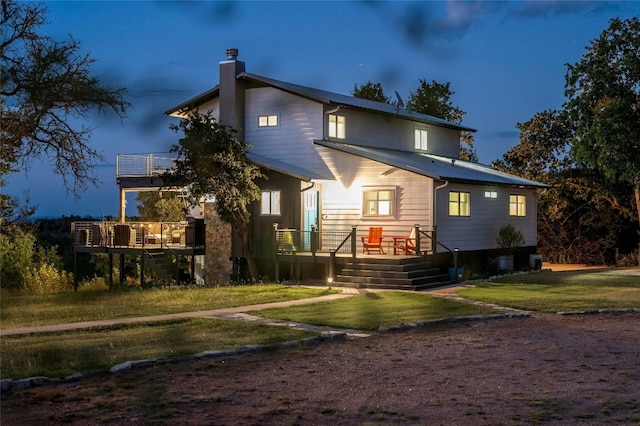 back of house at dusk featuring a balcony, a yard, a chimney, and a wooden deck