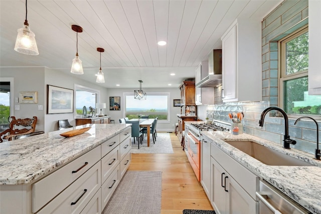 kitchen with a sink, white cabinetry, hanging light fixtures, appliances with stainless steel finishes, and wall chimney range hood