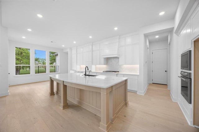kitchen with custom exhaust hood, a sink, light wood finished floors, stainless steel appliances, and decorative backsplash