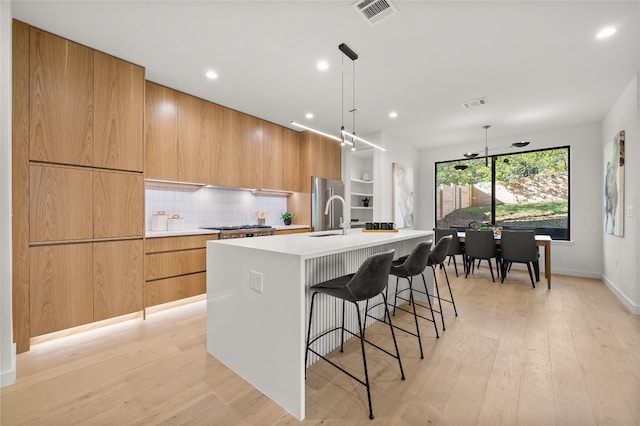 kitchen with hanging light fixtures, light hardwood / wood-style flooring, backsplash, stainless steel fridge, and a center island with sink