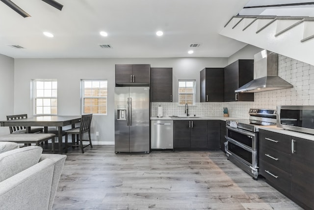 kitchen featuring decorative backsplash, stainless steel appliances, wall chimney range hood, and light hardwood / wood-style floors