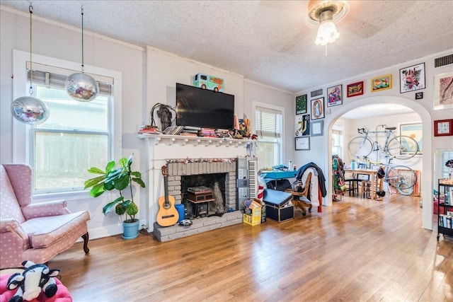 living room with a brick fireplace, a textured ceiling, ceiling fan, crown molding, and hardwood / wood-style flooring