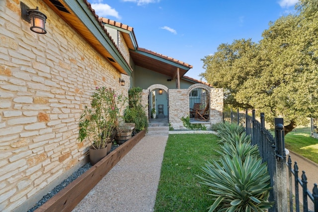 view of exterior entry with stucco siding, a lawn, a tile roof, stone siding, and fence