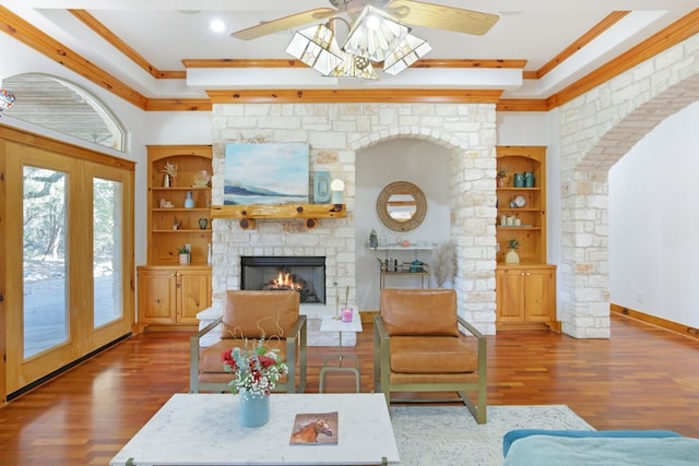 living room featuring built in shelves, ceiling fan, a stone fireplace, crown molding, and wood-type flooring