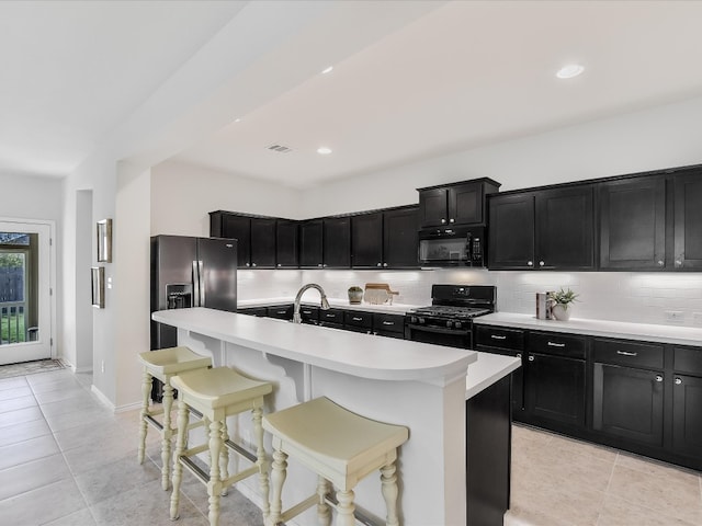 kitchen featuring decorative backsplash, light tile patterned flooring, black appliances, a breakfast bar area, and an island with sink