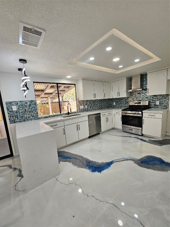 kitchen with appliances with stainless steel finishes, a tray ceiling, pendant lighting, and wall chimney range hood