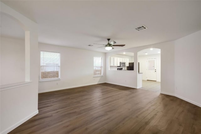 empty room with arched walkways, dark wood finished floors, visible vents, a ceiling fan, and baseboards