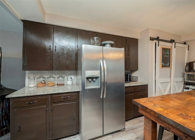 kitchen featuring dark brown cabinetry, light stone countertops, a barn door, stainless steel fridge with ice dispenser, and decorative backsplash