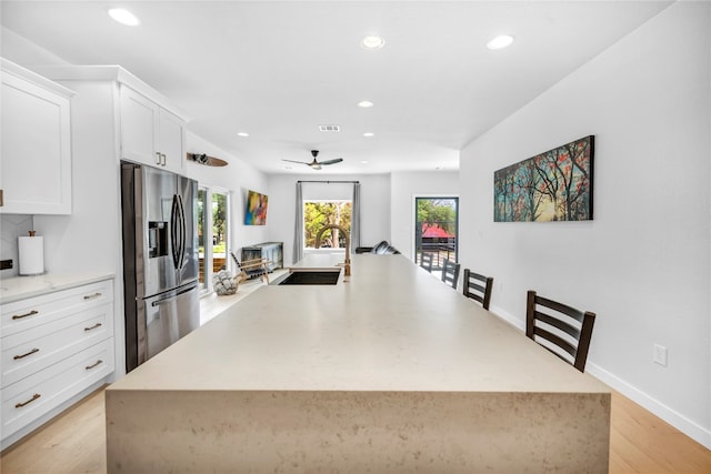 kitchen with ceiling fan, light hardwood / wood-style flooring, stainless steel fridge with ice dispenser, white cabinetry, and an island with sink