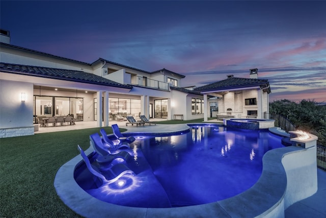 pool at dusk featuring a lawn, an outdoor stone fireplace, an in ground hot tub, and a patio