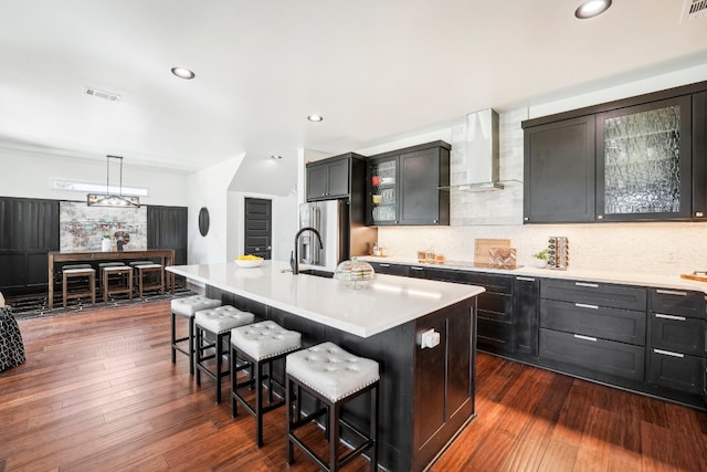 kitchen with high end fridge, a breakfast bar, an island with sink, and wall chimney range hood