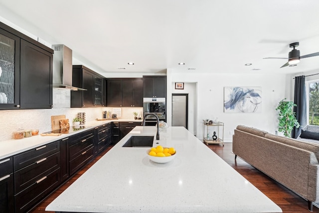 kitchen featuring wall chimney exhaust hood, sink, light stone counters, tasteful backsplash, and an island with sink