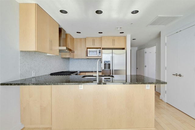 kitchen featuring kitchen peninsula, appliances with stainless steel finishes, dark stone counters, sink, and wall chimney range hood