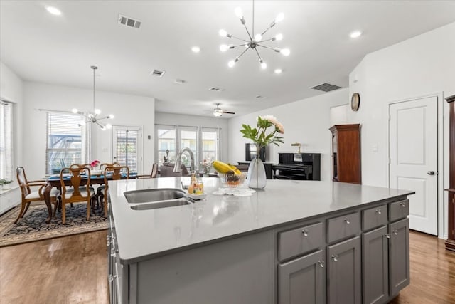 kitchen with sink, dark wood-type flooring, an island with sink, gray cabinets, and ceiling fan with notable chandelier