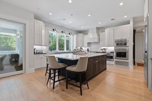 kitchen with white cabinets, a kitchen island with sink, stainless steel appliances, custom range hood, and light wood-type flooring