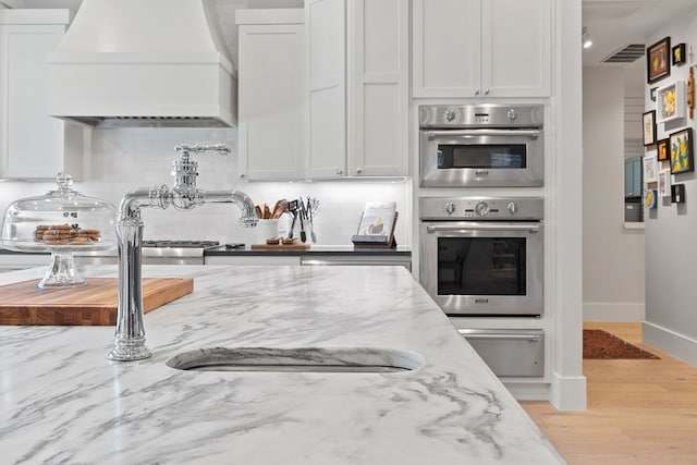 kitchen with light stone counters, white cabinetry, custom exhaust hood, and stainless steel double oven