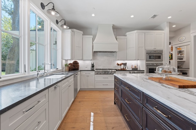 kitchen with sink, appliances with stainless steel finishes, dark stone countertops, custom range hood, and white cabinets