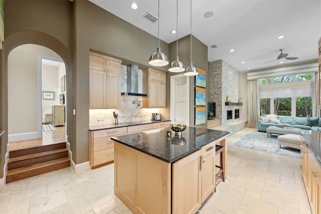 kitchen with light brown cabinets, a center island, wall chimney range hood, tasteful backsplash, and a stone fireplace