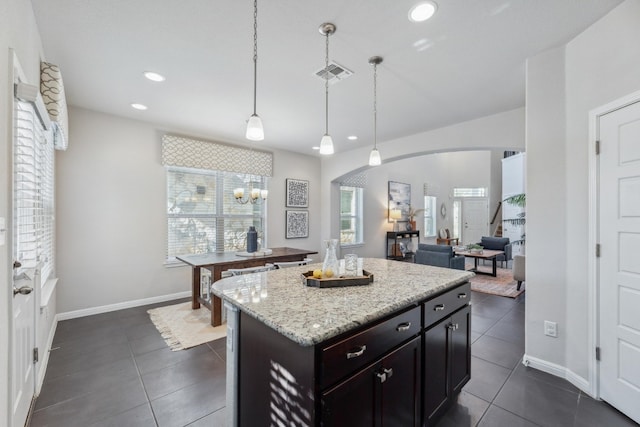 kitchen with dark tile patterned floors, dark brown cabinets, a kitchen island, pendant lighting, and light stone counters