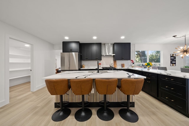 kitchen with wall chimney exhaust hood, a kitchen breakfast bar, light hardwood / wood-style flooring, stainless steel fridge, and a center island with sink